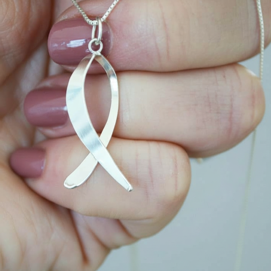 Silver ribbon ichthus pendant held between fingers against a neutral background